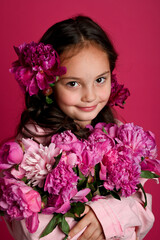 Naklejka premium A smiling girl with brown hair, adorned with pink flowers, holds a bouquet of vibrant pink peonies against a bright pink background.