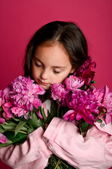 A young girl of Caucasian descent gently cradles a bouquet of vibrant pink peonies against a...