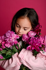 A young girl of Asian descent embraces a bouquet of vibrant pink peonies against a striking pink...