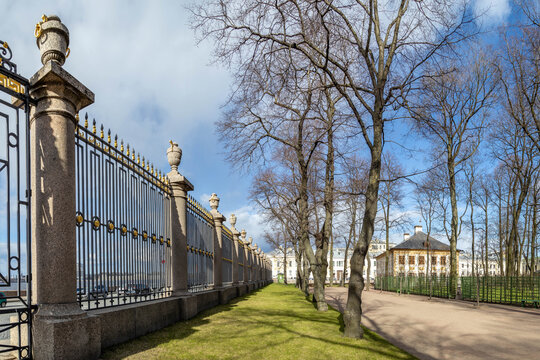 Classical cast-iron fence (1826) with gilded details borders Summer Garden's bare-tree alleys. Iconic Neva embankment landmark in spring sunlight. St. Petersburg, Russia. 