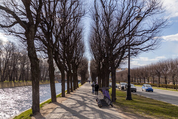 Spring sunlight on Moyka River promenade with parked scooters, tree shadows and reflections in water under blue sky. Saint Petersburg, Russia

