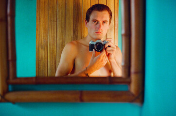 A shirtless young man holds a vintage 35mm film camera, capturing his reflection in a stylish mirror. The setting combines natural textures with vibrant colours, creating a striking portrait.