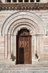 Medieval sculpted side portal of Foligno Cathedral, Umbria, Italy. Romanesque‑Gothic arch with pink marble bands, intricate carvings and sun‑lit wooden door