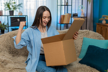 Young happy woman at home happily unpacking delivery box, feeling pleasantly surprised pulling out...