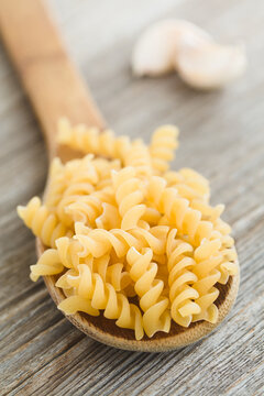 Raw fusilli or rotini pasta on wooden spoon, with garlic cloves in the back (Selective Focus, Focus one third into the image)