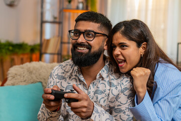 Indian couple man and woman sitting on home sofa with game controller and popcorn, laughing and...