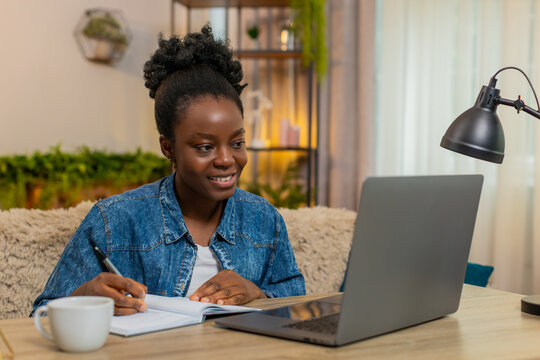 African American young woman studies online at laptop while sitting at table on home sofa. Black girl takes notes in notebook looking focused, attentive, and fully engaged in distance learning process