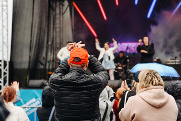 music festival in city park, crowd of people backview, rock group performing on stage, green trees and summer day