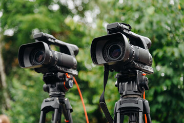 two large black video cameras on tripod outdoors, television work
