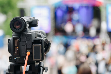 large black video camera on tripod outdoors, shooting music festival and crowd of people outdoors, television work