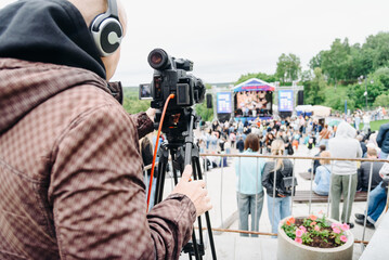 large black video camera on tripod outdoors, operator videographer shooting music festival and crowd of people outdoors, television work