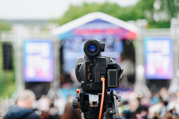 large black video camera on tripod outdoors, shooting music festival and crowd of people outdoors, television work