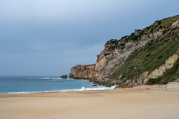 Fototapeta premium Scenic view of dramatic coastal cliffs meeting the ocean beside a wide empty beach with soft waves and cloudy sky.