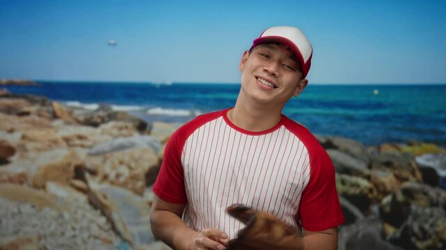 Young man in a baseball uniform playfully tosses a ball on a picturesque seaside setting with a clear blue ocean and rocky beach backdrop.