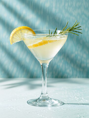 Stylish cocktail in a classic martini glass, garnished with fresh rosemary and lemon slice, illuminated by natural light with palm shadow accents on a pastel backdrop.
