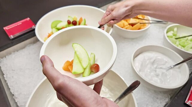 Person serves sliced fruits into bowl from ice-filled hotel buffet station with yogurt and fresh options, healthy start