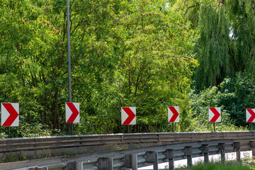 Curved road with multiple red and white chevron direction signs mounted along a guardrail in a green forest setting. Traffic safety symbols warning of a sharp right turn ahead. Bright summer day,