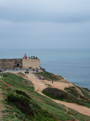 People walk on a coastal path and gather near the red lighthouse on a cliff edge overlooking the Atlantic Ocean under cloudy skies.