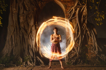 Female performer standing in front of a doorway covered in tree roots fire dancing, Thailand