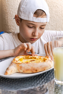 Teenage boy in a baseball cap sitting in a street cafe eating a Traditional Adjarian khachapuri with Lagidze water