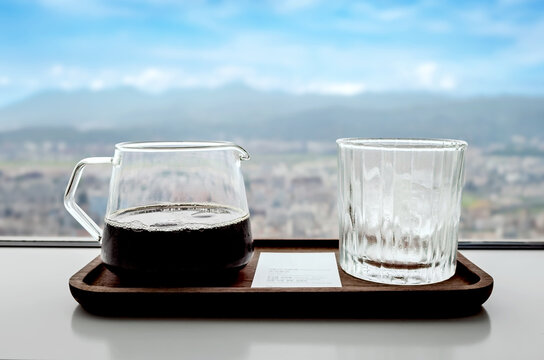 Close-up of a transparent glass jug of black Coffee and a glass of ice cubes on a tray by a Window Overlooking a city