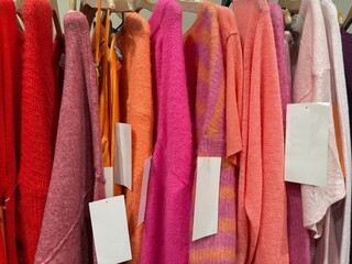 Close-up of assorted pink, orange and red clothes hanging on a clothes rail 
for sale with blank tags in a shop