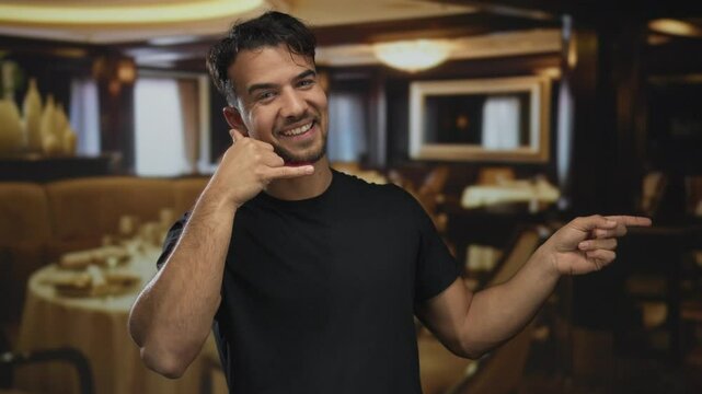Young hispanic man making a call gesture and smiling indoors at a cozy restaurant with elegant decor and ambient lighting, exuding a friendly and lively atmosphere.