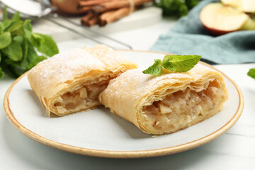 Pieces of tasty apple strudel with powdered sugar, mint, fruit and cinnamon on white marble table, closeup