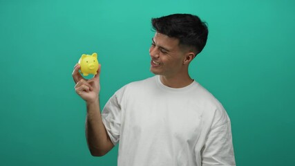 Young man holding yellow piggy bank smiling against green background showing happiness and prosperity in financial savings