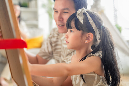 Happy Asian family spending quality time together at home. Father and daughter drawing on the floor with colored pens while mother works on a digital tablet smartphone on the sofa. Warm, modern living