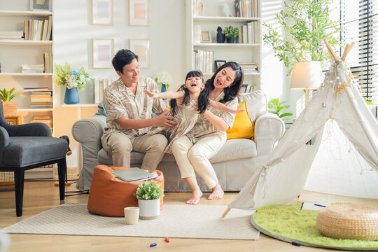 Happy Asian family enjoying quality time in their modern living room. Cheerful young parents playfully carry and bond with their daughter while laughing together on the sofa, creating joyful moments a