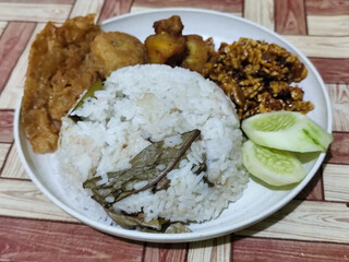 Complete Indonesian uduk rice served with tempe orek, egg, fried chicken, tofu fritters and cucumber slices on a white plate with a brown wooden patterned tablecloth in the background.