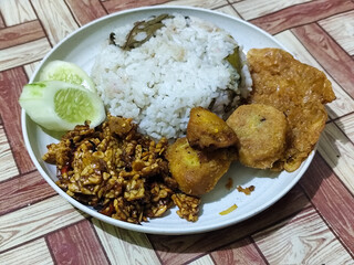 Complete Indonesian uduk rice served with tempe orek, egg, fried chicken, tofu fritters and cucumber slices on a white plate with a brown wooden patterned tablecloth in the background.