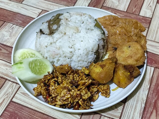 Complete Indonesian uduk rice served with tempe orek, egg, fried chicken, tofu fritters and cucumber slices on a white plate with a brown wooden patterned tablecloth in the background.