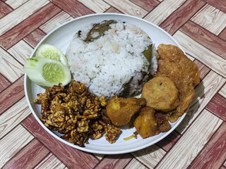 Complete Indonesian uduk rice served with tempe orek, egg, fried chicken, tofu fritters and cucumber slices on a white plate with a brown wooden patterned tablecloth in the background.