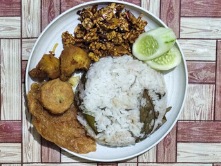 Complete Indonesian uduk rice served with tempe orek, egg, fried chicken, tofu fritters and cucumber slices on a white plate with a brown wooden patterned tablecloth in the background.