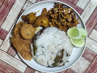 Complete Indonesian uduk rice served with tempe orek, egg, fried chicken, tofu fritters and cucumber slices on a white plate with a brown wooden patterned tablecloth in the background.