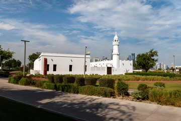 Traditional white mosque with a minaret stands in contrast to modern skyscrapers of West Bay in Doha, Qatar. Green park and blue sky create a peaceful urban landscape.
