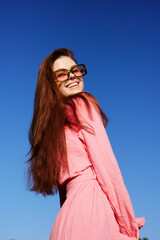 Young woman with long red hair smiling in a pink dress under a blue sky, showcasing vibrant summer...