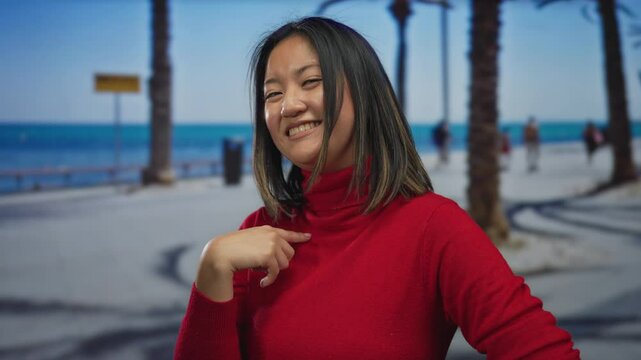 Young woman in red sweater pointing to herself while smiling at the seaside promenade with palm trees and ocean in the background.