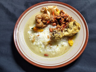 Bubur ayam. A warm Indonesian dish of chicken porridge with savory yellow sauce, chicken pieces, peanuts and orek tempeh served completely on a brown plate.
