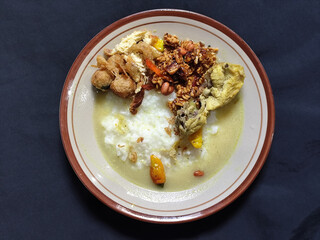 Bubur ayam. A warm Indonesian dish of chicken porridge with savory yellow sauce, chicken pieces, peanuts and orek tempeh served completely on a brown plate.