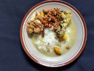 Bubur ayam. A warm Indonesian dish of chicken porridge with savory yellow sauce, chicken pieces, peanuts and orek tempeh served completely on a brown plate.
