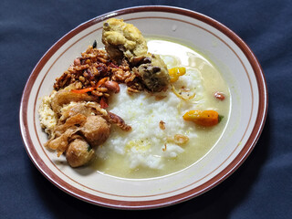 Bubur ayam. A warm Indonesian dish of chicken porridge with savory yellow sauce, chicken pieces, peanuts and orek tempeh served completely on a brown plate.