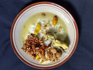 Bubur ayam. A warm Indonesian dish of chicken porridge with savory yellow sauce, chicken pieces, peanuts and orek tempeh served completely on a brown plate.