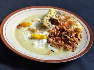 Bubur ayam. A warm Indonesian dish of chicken porridge with savory yellow sauce, chicken pieces, peanuts and orek tempeh served completely on a brown plate.