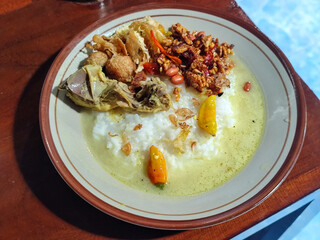 Bubur ayam. A warm Indonesian dish of chicken porridge with savory yellow sauce, chicken pieces, peanuts and orek tempeh served completely on a brown plate.
