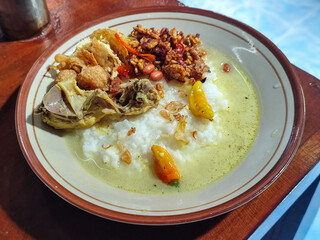 Bubur ayam. A warm Indonesian dish of chicken porridge with savory yellow sauce, chicken pieces, peanuts and orek tempeh served completely on a brown plate.