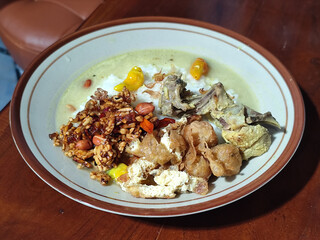 Bubur ayam. A warm Indonesian dish of chicken porridge with savory yellow sauce, chicken pieces, peanuts and orek tempeh served completely on a brown plate.