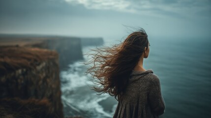 A contemplative woman stands by a cliff, gazing at the ocean waves under a moody sky, feeling the wind in her hair.